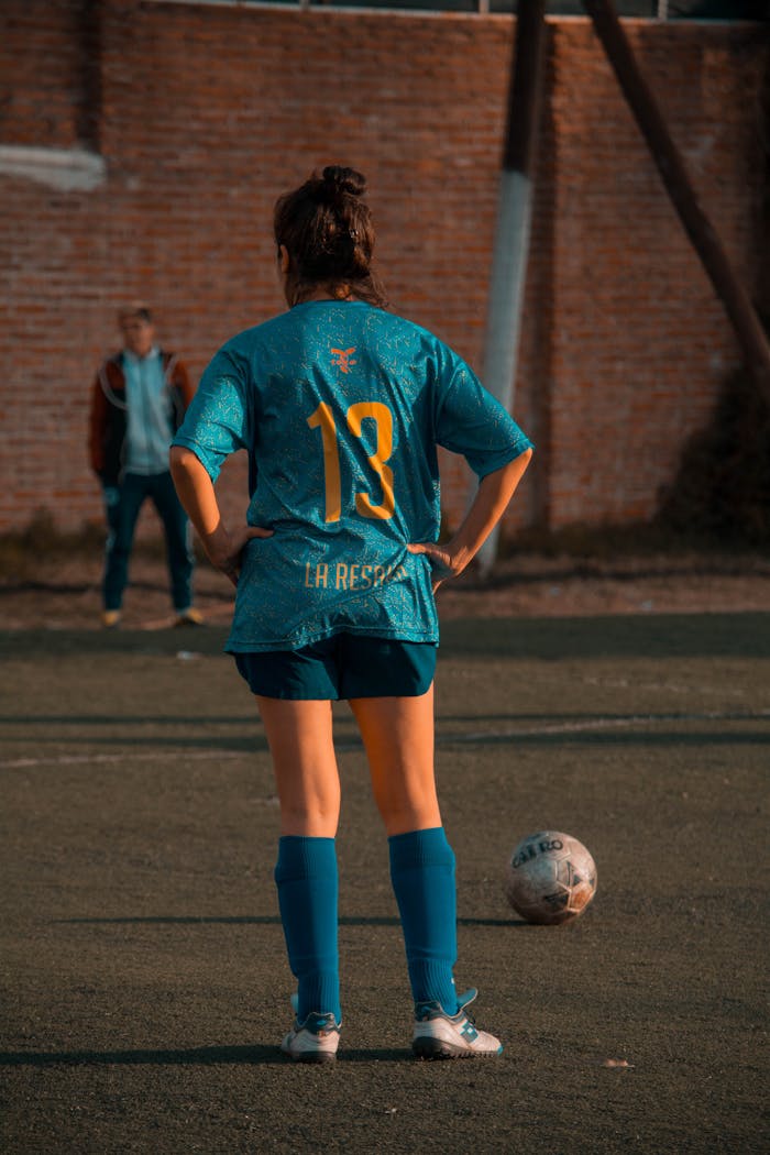 A female soccer player stands on the field, preparing for a game under a warm setting sun.